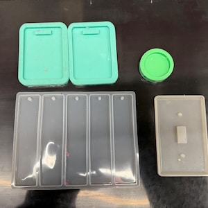 May include: An assortment of silicone molds in various shapes and colors. Includes two rectangular teal molds, a round green mold, a rectangular mold with multiple slots, and a light gray light switch cover mold. Ideal for crafting projects.