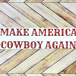 May include: A white rectangular sticker with the phrase "MAKE AMERICA COWBOY AGAIN" in red, stylized text. The sticker is set against a light brown, chevron-patterned wooden background.