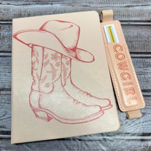 May include: A light pink notebook featuring a red illustration of a cowboy hat and boots. A matching pink pen holder with the word "COWGIRL" is attached. The notebook and pen holder are on a wooden surface.