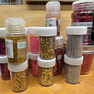 Puede incluir: Una colección de botes de purpurina en varios colores, incluyendo dorado, plateado, rosa y rojo. Los botes son transparentes con tapas blancas y están colocados sobre una superficie de madera. Algunos botes tienen la etiqueta "Craft glitter ultra".