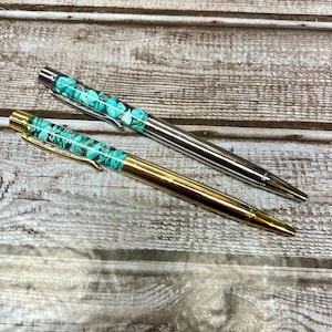 May include: Two pens with turquoise blue and silver accents. The pens have a clear barrel filled with turquoise blue stones. One pen has a silver barrel and the other has a gold barrel.