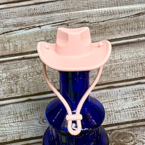May include: A pink cowboy hat bottle stopper with a blue glass bottle. The bottle stopper has a strap that goes around the neck of the bottle.