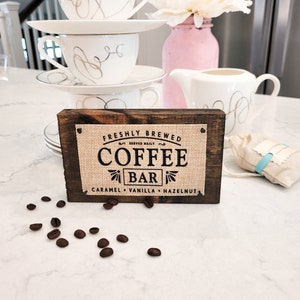 May include: A wooden sign with a burlap background reads "Freshly Brewed Coffee Bar Served Daily Caramel Vanilla Hazelnut". The sign is sitting on a white countertop with coffee beans scattered around it.