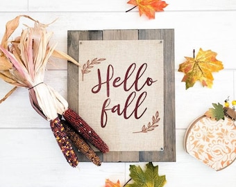 May include: A burlap sign with the text "Hello Fall" in burgundy script. The sign is framed with a rustic wood frame. The sign is surrounded by fall decor, including corn husks, Indian corn, and maple leaves.