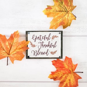 May include: A white wooden sign with the words "grateful thankful blessed" in maroon script with small brown leaf accents. The sign is surrounded by three orange and yellow fall leaves.