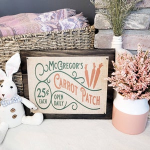 May include: A rustic wooden sign with a burlap background featuring the text "McGregor's Carrot Patch" in green and orange lettering. The sign also includes the text "25¢ Each Open Daily".