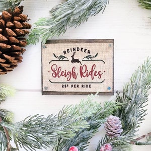 May include: A rustic wooden sign with burlap fabric and a red and black design. The sign reads "Reindeer Sleigh Rides 25¢ Per Ride".