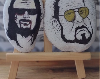 May include: Two painted rocks on a wooden easel. One rock features a portrait with black hair, beard, and sunglasses. The other rock has a portrait with yellow sunglasses and a beard. The background is a white and black pattern.