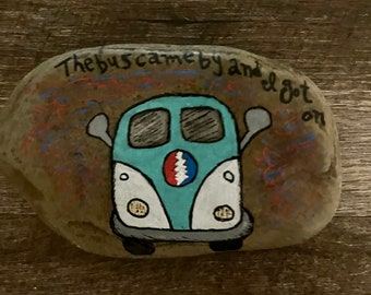 May include: Hand-painted rock art featuring a light blue and white VW bus with a red, white, and blue emblem. The text "The bus came by and I got on" is written above the bus in black and red.