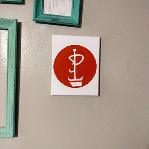 May include: A small, white canvas art piece featuring a bold, red circle with a white, stylized symbol. The symbol appears to be a combination of a capital P and a stylized flame or lightbulb. The artwork is mounted on a neutral-colored wall.