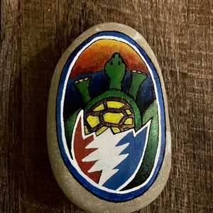 May include: A hand-painted, oval-shaped rock featuring a green turtle design within a blue and white emblem. The emblem is set against a backdrop of orange and blue. The rock is on a wooden surface.