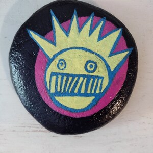 May include: A hand-painted, black rock featuring a whimsical face design. The face has a yellow crown, blue eyes and mouth, and a pink circle background. The rock is a unique piece of art.
