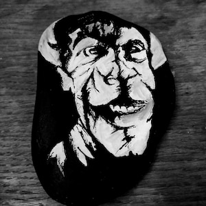 May include: Black and white painted rock art featuring a caricature of a smiling face. The artwork is on a smooth, dark-colored stone, with the face rendered in bold white brushstrokes against a black background. The rock is displayed on a wooden surface.