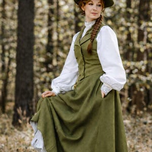 Op de afbeelding: Een olijfgroene jurk in Victoriaanse stijl met een bijpassend vest en lange witte mouwen. De jurk heeft een hoge kraag en kanten manchetten. Een bruine hoed met veren maakt de outfit compleet. De outfit wordt buiten in een bos gedragen.