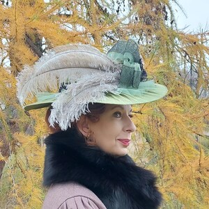 May include: A light green hat with a wide brim, adorned with large, fluffy feathers in shades of gray and beige. The hat features a dark green ribbon and is set against a backdrop of fall foliage. A black fur collar is worn.