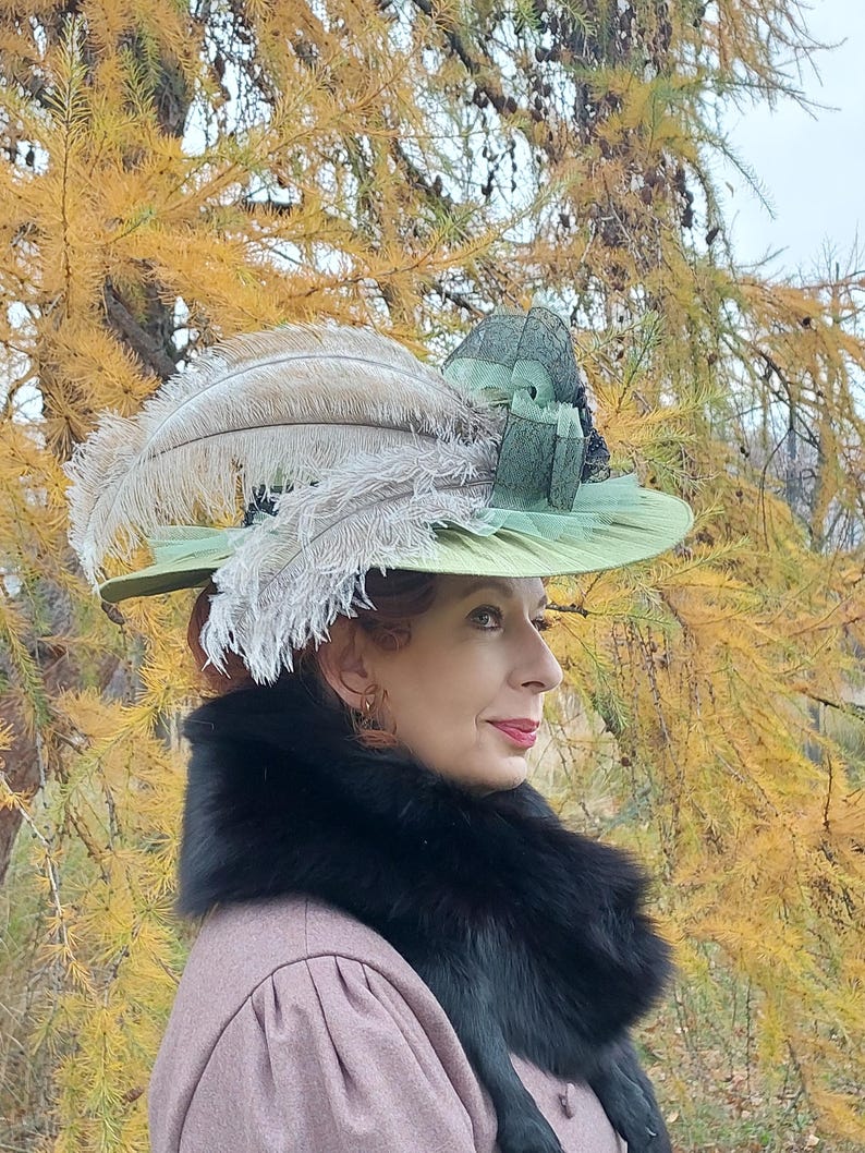 May include: A light green hat with a wide brim, adorned with large, fluffy feathers in shades of gray and beige. The hat features a dark green ribbon and is set against a backdrop of fall foliage. A black fur collar is worn.