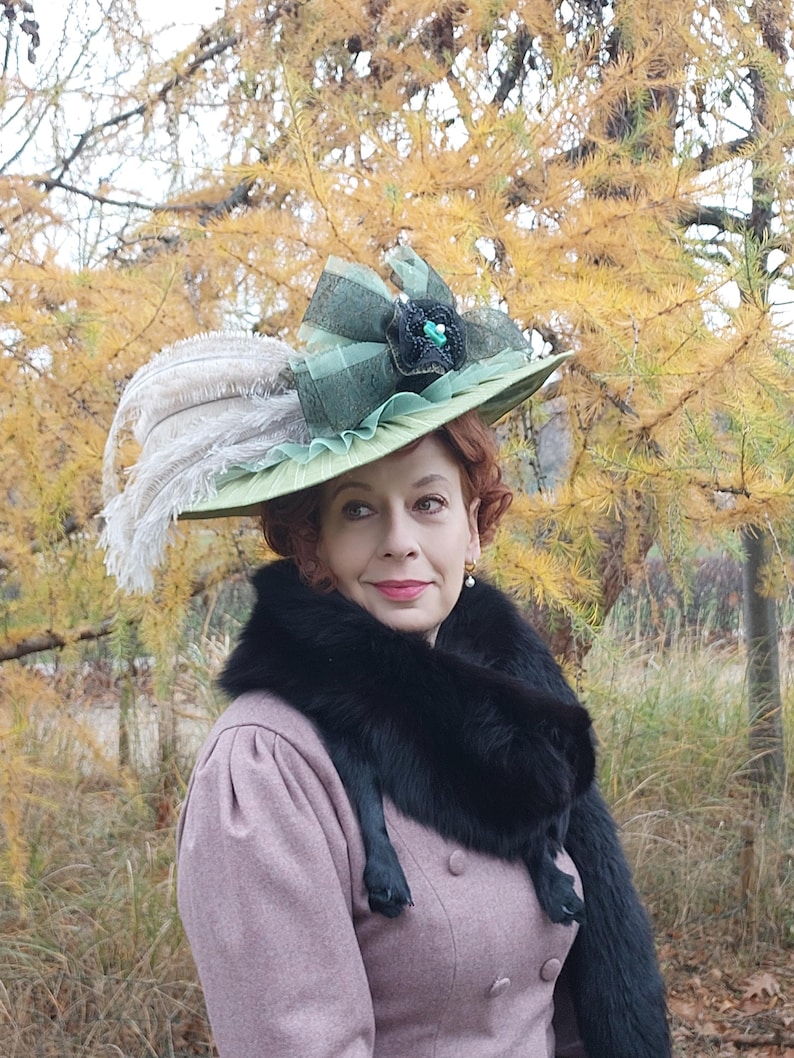 May include: A light green hat with a wide brim, adorned with a large green bow, black embellishments, and white feathers. The hat is worn with a pink coat and a black fur stole.