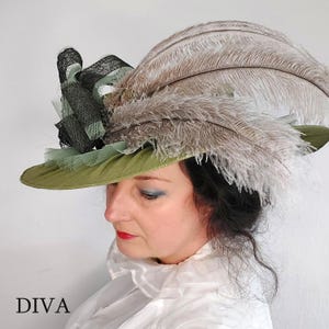 May include: An olive green hat with a wide brim, adorned with large, light brown feathers and black lace detailing. The hat has a decorative arrangement of black and light green fabric. The word "DIVA" is printed in black at the bottom.