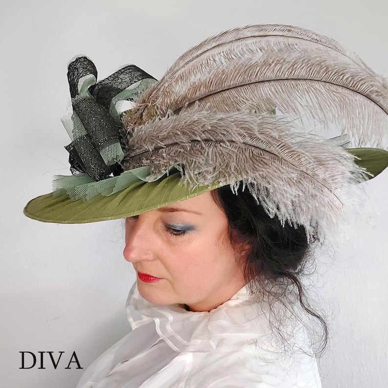May include: An olive green hat with a wide brim, adorned with large, light brown feathers and black lace detailing. The hat has a decorative arrangement of black and light green fabric. The word "DIVA" is printed in black at the bottom.