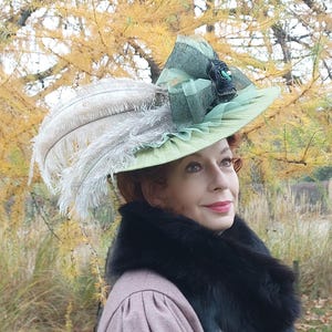 May include: A light green hat with a wide brim, adorned with white feathers and a decorative bow. The hat features a black and green embellishment. The person is wearing a pink coat and a black fur collar.