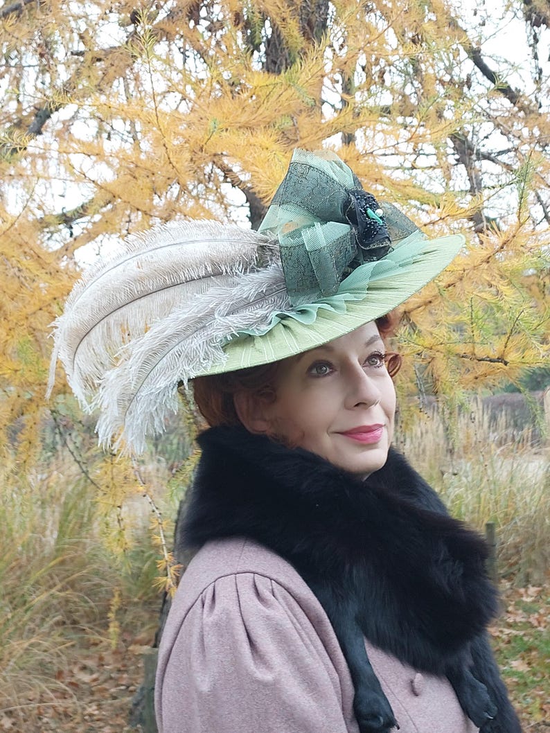 May include: A light green hat with a wide brim, adorned with white feathers and a decorative bow. The hat features a black and green embellishment. The person is wearing a pink coat and a black fur collar.