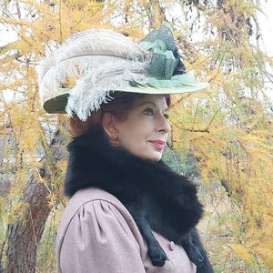 May include: A vintage-style hat with a wide brim, adorned with large, fluffy white feathers and a green ribbon. The hat is set against a backdrop of fall foliage. The woman is wearing a pink dress and a black fur collar.