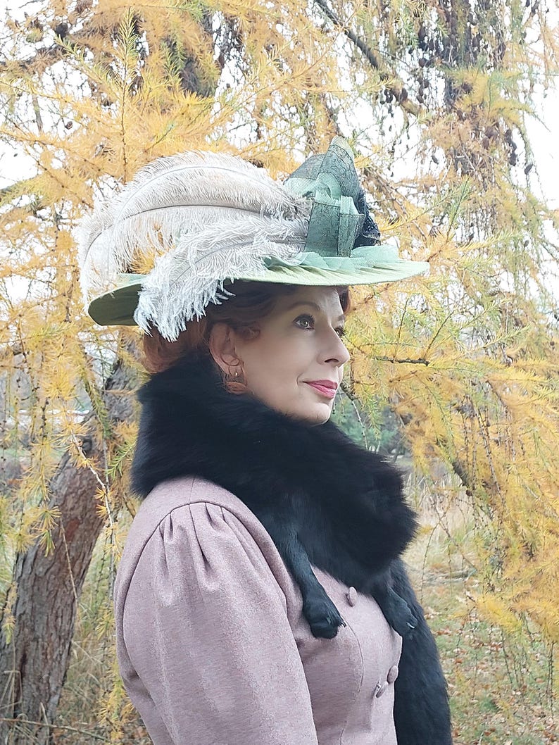 May include: A vintage-style hat with a wide brim, adorned with large, fluffy white feathers and a green ribbon. The hat is set against a backdrop of fall foliage. The woman is wearing a pink dress and a black fur collar.