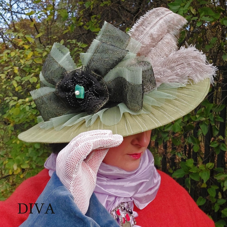 May include: A wide-brimmed, olive-green hat with a decorative black flower, green netting, and tan feathers. The hat has a large, decorative bow with dark green and black lace. The word "DIVA" is printed in white on the lower left.