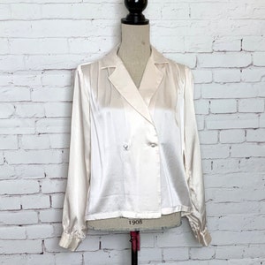May include: A long-sleeved, double-breasted blouse in a pale cream satin fabric. This garment features a notched lapel collar, two small matching buttons on the front closure, and cuffed sleeves, displayed on a mannequin torso.