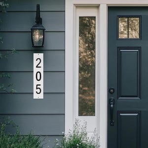 May include: A white rectangular address sign with the number "205" in black, mounted on a blue-gray house exterior. A black lantern hangs above the sign. The door is dark blue-green.