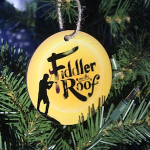 May include: A round, yellow Christmas ornament with the words "Fiddler on the Roof" in black script. A black silhouette of a fiddler is on the left. The ornament hangs from a twine string on a green Christmas tree.