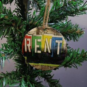 May include: A round Christmas ornament with the word "RENT" in red, green, blue, and yellow block letters. The ornament has a distressed, graffiti-style design and is suspended by a natural twine string. The ornament is hanging on a green Christmas tree.