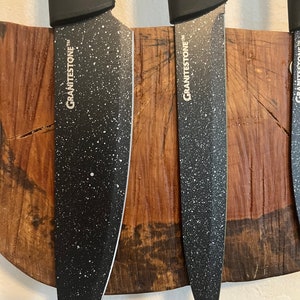May include: Three black kitchen knives with white speckled blades are mounted on a wooden magnetic strip. The knives have the word "GRANITESTONE" printed on the side.