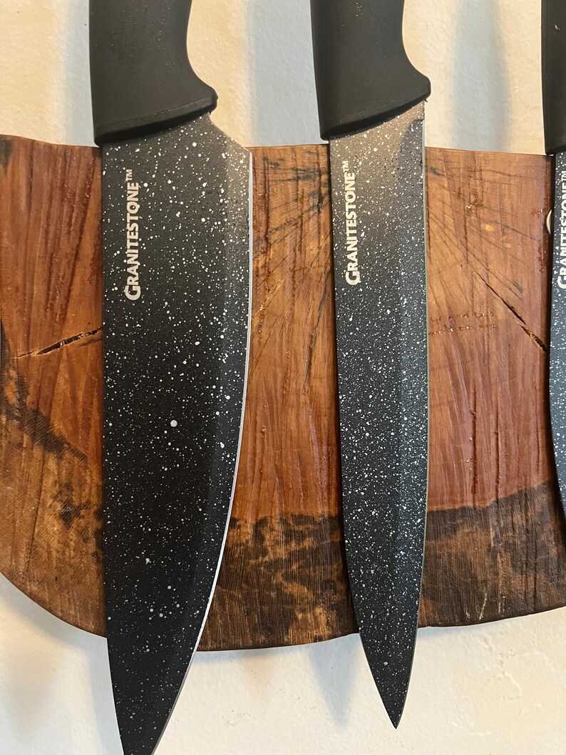 May include: Three black kitchen knives with white speckled blades are mounted on a wooden magnetic strip. The knives have the word "GRANITESTONE" printed on the side.