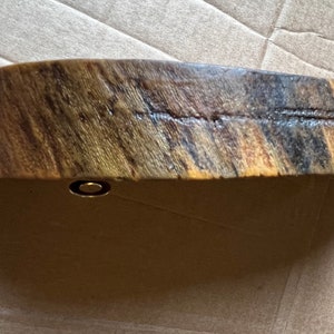 May include: A curved piece of wood with a natural wood grain pattern. The wood is a mix of brown, black, and yellow tones. A small gold metal piece is attached to the bottom of the wood.