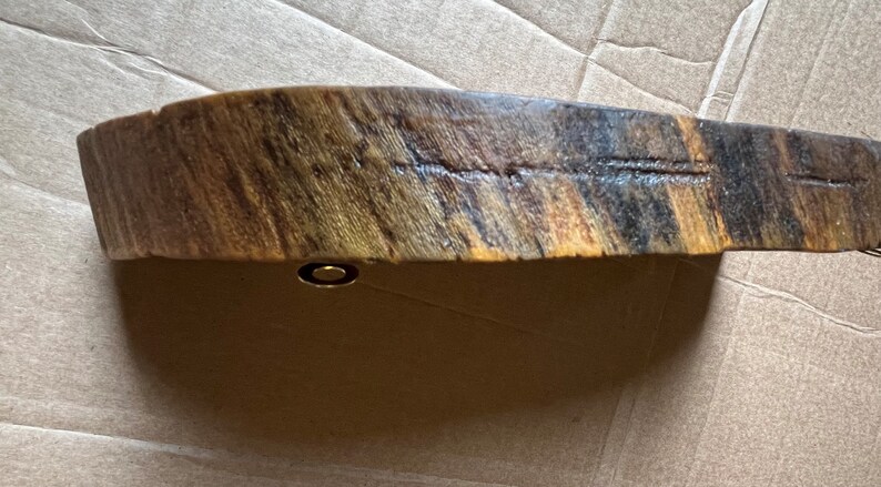 May include: A curved piece of wood with a natural wood grain pattern. The wood is a mix of brown, black, and yellow tones. A small gold metal piece is attached to the bottom of the wood.