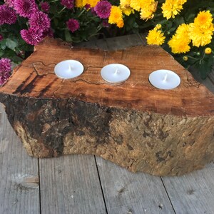 May include: A rustic wooden candle holder with three white tea lights. The holder is made from a natural tree trunk slice with a rough bark texture.