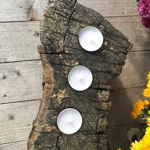 May include: A rustic wooden candle holder with three white tea lights. The holder is made from a natural piece of wood with a rough, textured surface.