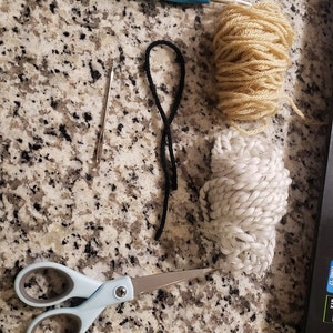 Peut inclure: Gros plan sur des fournitures d'artisanat, notamment un crochet bleu, une pelote de laine blanche, une pelote de laine beige, un cordon noir, une aiguille argent&eacute;e et une paire de ciseaux bleus.