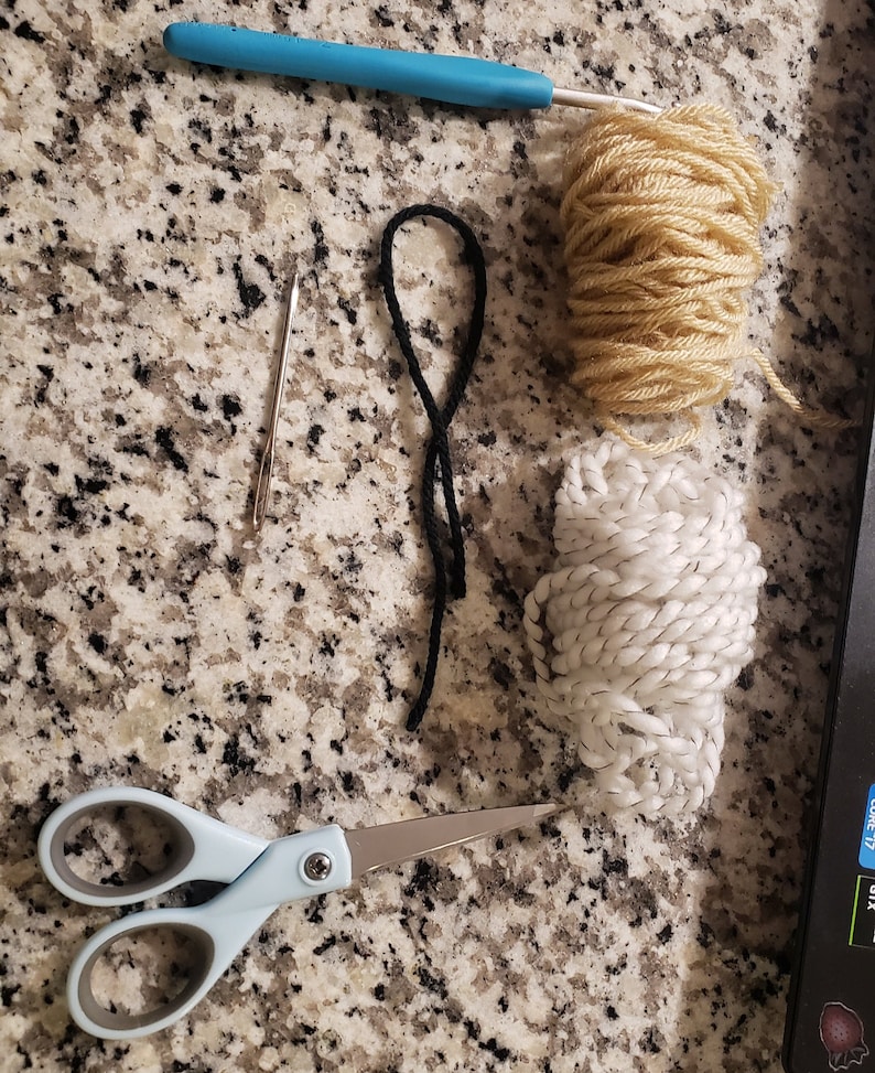 Peut inclure: Gros plan sur des fournitures d'artisanat, notamment un crochet bleu, une pelote de laine blanche, une pelote de laine beige, un cordon noir, une aiguille argent&eacute;e et une paire de ciseaux bleus.