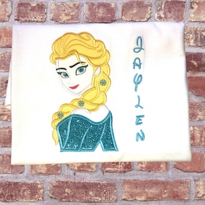 K&ouml;nnte beinhalten: Wei&szlig;es T-Shirt mit einem gestickten Bild von Elsa aus Frozen und dem Namen "Jaylen" in blauen Buchstaben.