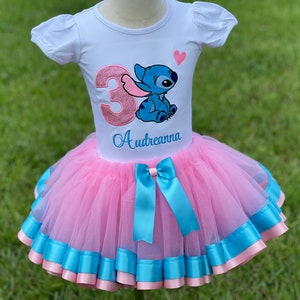 May include: A pink tulle tutu skirt with blue ribbon trim and a matching blue bow. A white t-shirt with a pink sparkly number 3 and a blue Stitch character with a pink heart. The shirt is personalized with the name "Audreanna".
