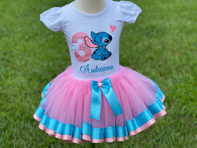 May include: A pink tulle tutu skirt with blue ribbon trim and a matching blue bow. A white t-shirt with a pink sparkly number 3 and a blue Stitch character with a pink heart. The shirt is personalized with the name "Audreanna".