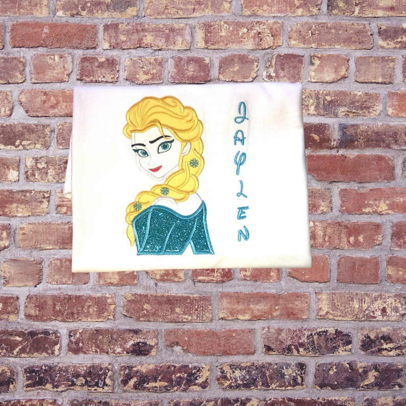K&ouml;nnte beinhalten: Wei&szlig;es T-Shirt mit einem gestickten Bild von Elsa aus Frozen und dem Namen "Jaylen" in blauen Buchstaben.