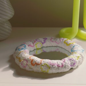 May include: A white, round, decorative mirror with a raised, textured surface. The mirror is decorated with colorful, painted flowers and stars. The mirror is sitting on a white surface.