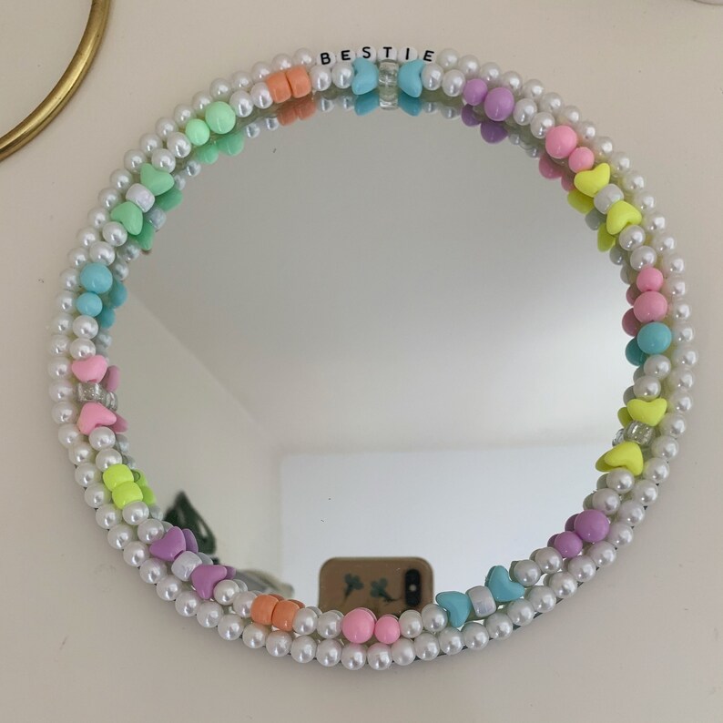 Puede incluir: Un espejo redondo con un marco de cuentas de perlas blancas. Las cuentas est&aacute;n intercaladas con cuentas de colores pastel, incluyendo rosa, azul, verde y amarillo. La palabra "BESTIE" est&aacute; escrita en letras negras en la parte superior del espejo.