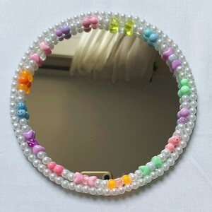 Puede incluir: Un espejo redondo con un marco de cuentas de perlas blancas. Las cuentas est&aacute;n decoradas con abalorios de pl&aacute;stico de colores, incluyendo corazones, caritas sonrientes y lazos.