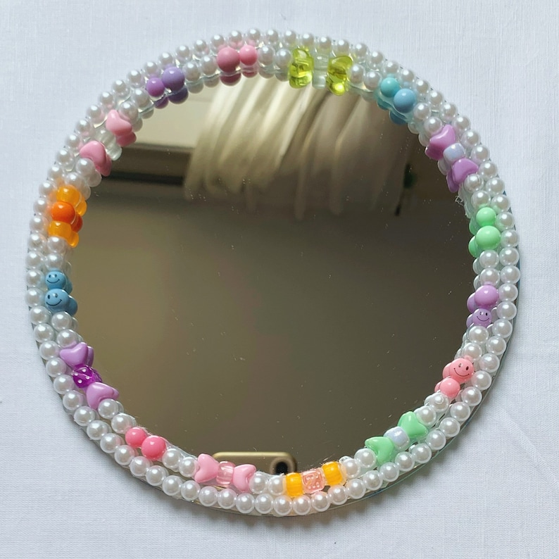 Puede incluir: Un espejo redondo con un marco de cuentas de perlas blancas. Las cuentas est&aacute;n decoradas con abalorios de pl&aacute;stico de colores, incluyendo corazones, caritas sonrientes y lazos.