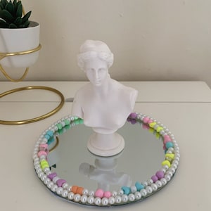 Puede incluir: Un busto de yeso blanco de una mujer con una bandeja de espejo decorada con una guirnalda de cuentas de colores.