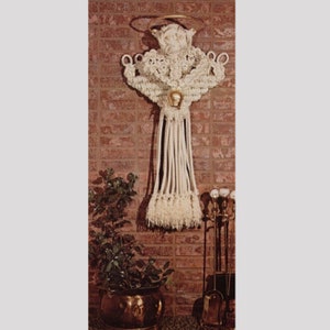 Op de afbeelding: Een macramé engel wandkleed met een gouden halo en een goudkleurig gezicht. De engel is gemaakt van gebroken wit touw en hangt tegen een rode bakstenen muur. Een plant in een pot en haardgereedschap zijn zichtbaar op de voorgrond.